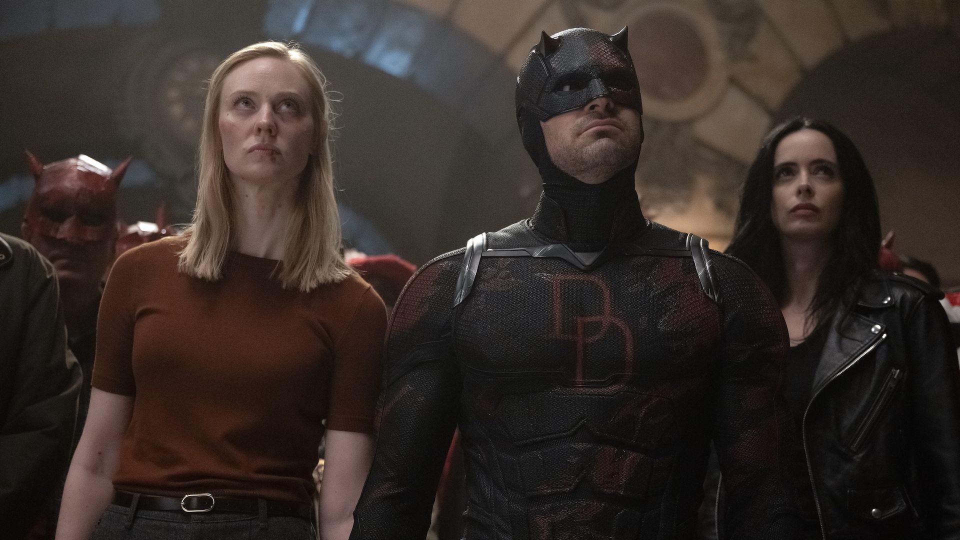 Deborah Ann Woll's Karen wearing maroon shirt standing next to Charlie Cox's Daredevil in black uniform, with Krysten Ritter's Jessica Jones in leather jacket in Daredevil: Born Again Season 2