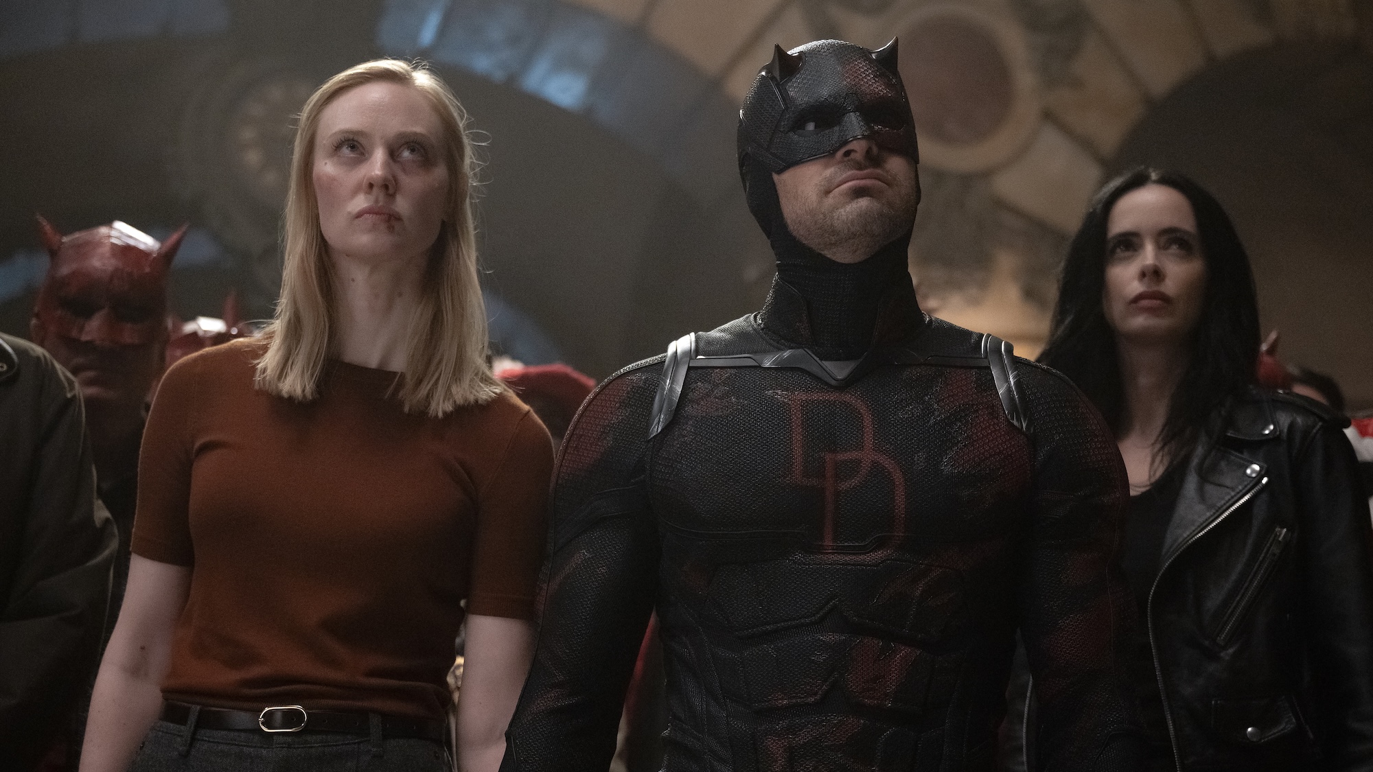 Deborah Ann Woll's Karen wearing maroon shirt standing next to Charlie Cox's Daredevil in black uniform, with Krysten Ritter's Jessica Jones in leather jacket in Daredevil: Born Again Season 2