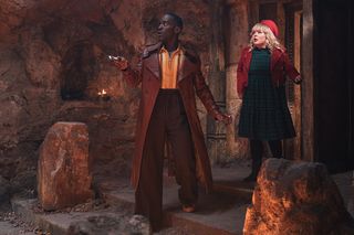 The Doctor (Ncuti Gatwa) and Joy (Nicola Coughlan) walk through a door into a large stone room. The Doctor is walking down concrete steps, and there are waist-height stone pillars on either side of the steps. The Doctor is using his sonic screwdriver like a torch.