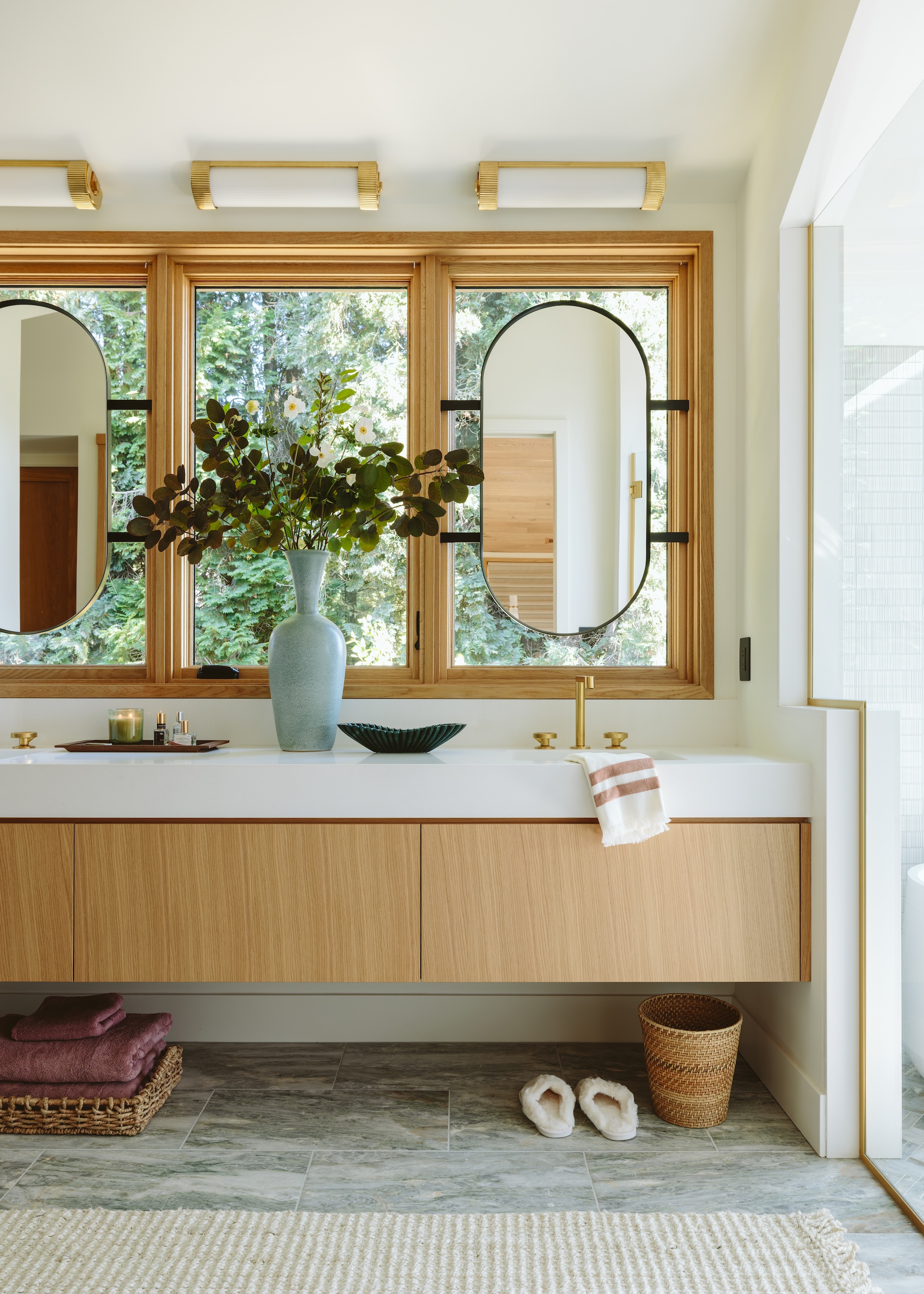 A bathroom with double mirrors on top of a window and two sinks with a vase and plant between it