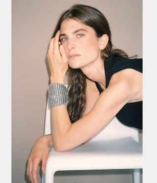 Image of Maria Sole Ferragamo, leaning on a small white chair, wearing a black sleeveless dress, hair hung down, modelling a thick band silver, sparkly bracelet, beige backdrop