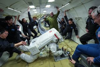 a person in a bulky spacesuit floats in an aircraft fuselage surrounded by people in blue flight suits