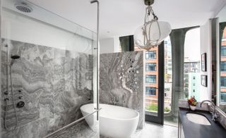 A bathroom with grey marble walls, an oval tub, a clear glass shower area and a black vanity.