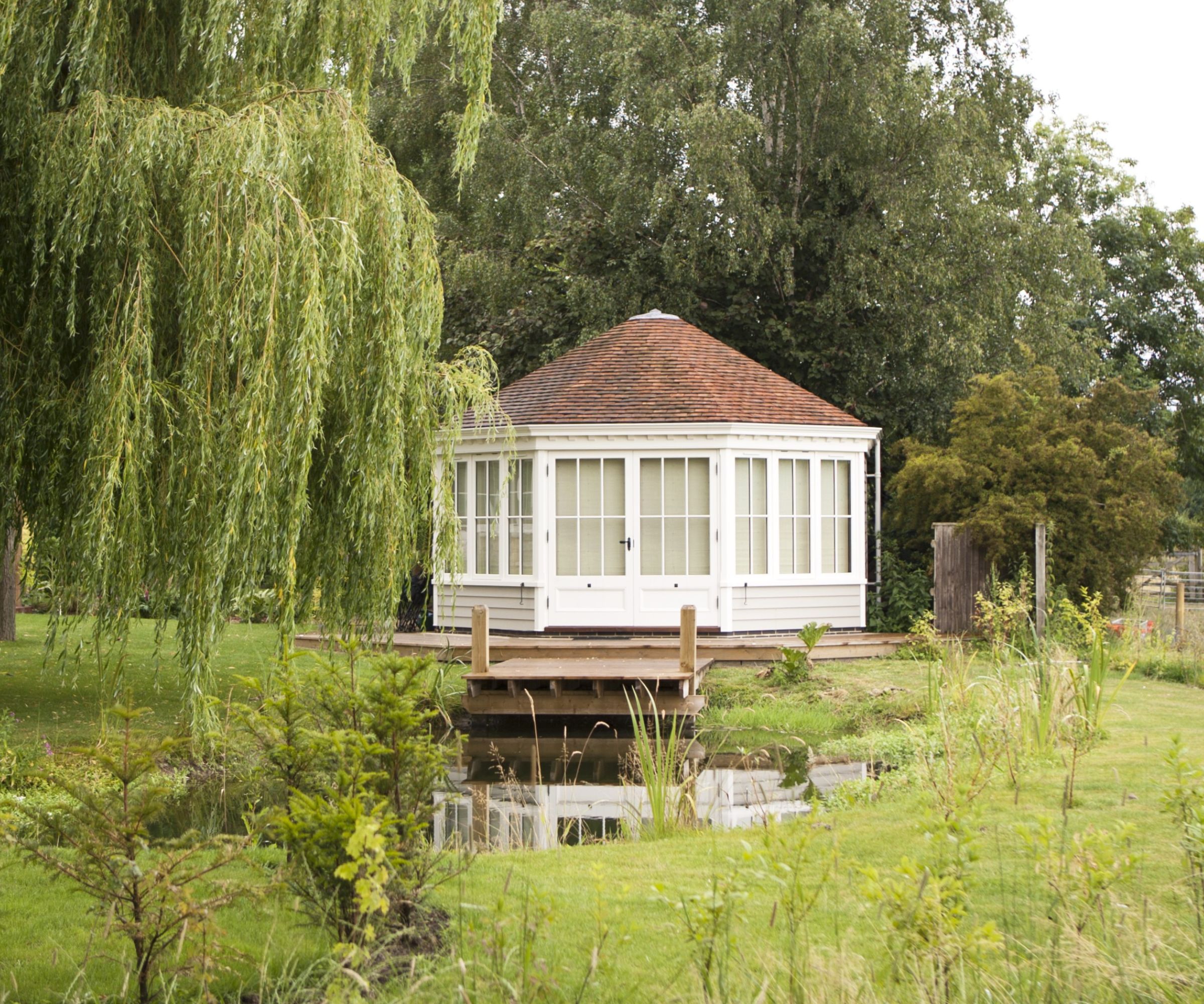 White octagonal garden pavilion with tiled roof beside a pond, willow tree draping over lawn in foreground.