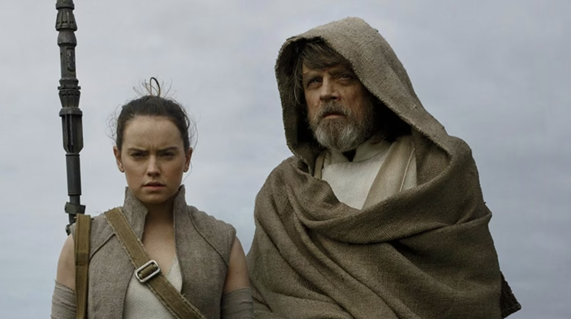 Rey and a hooded, bearded Luke Skywalker stand side by side with serious expressions under an overcast sky.