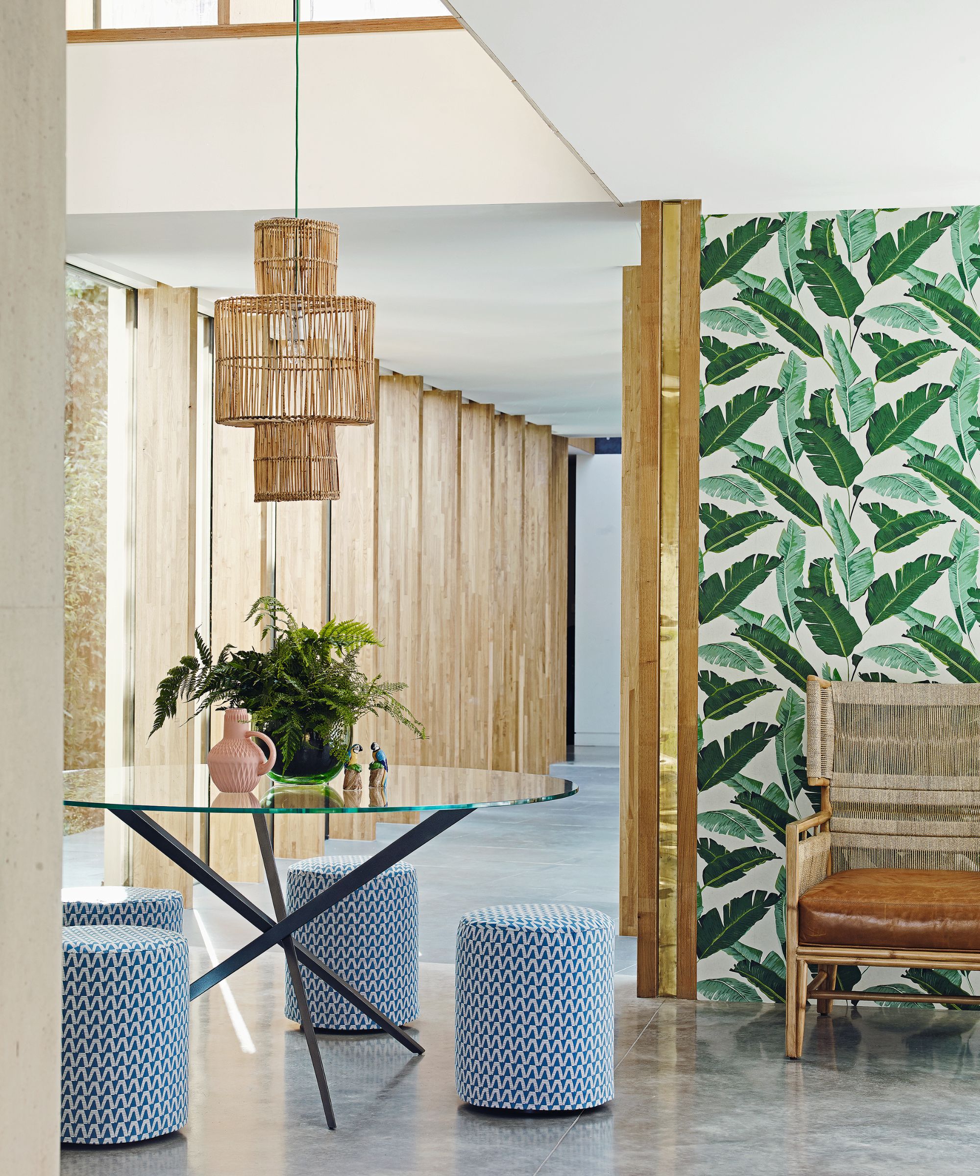 A bright and modern green and wood modern layered living space with a green wallpapered statement wall, glass table with leafy plant, blue patterned stools and a wooden bench with leather cushion. Above is a hanging light with rattan lampshade.