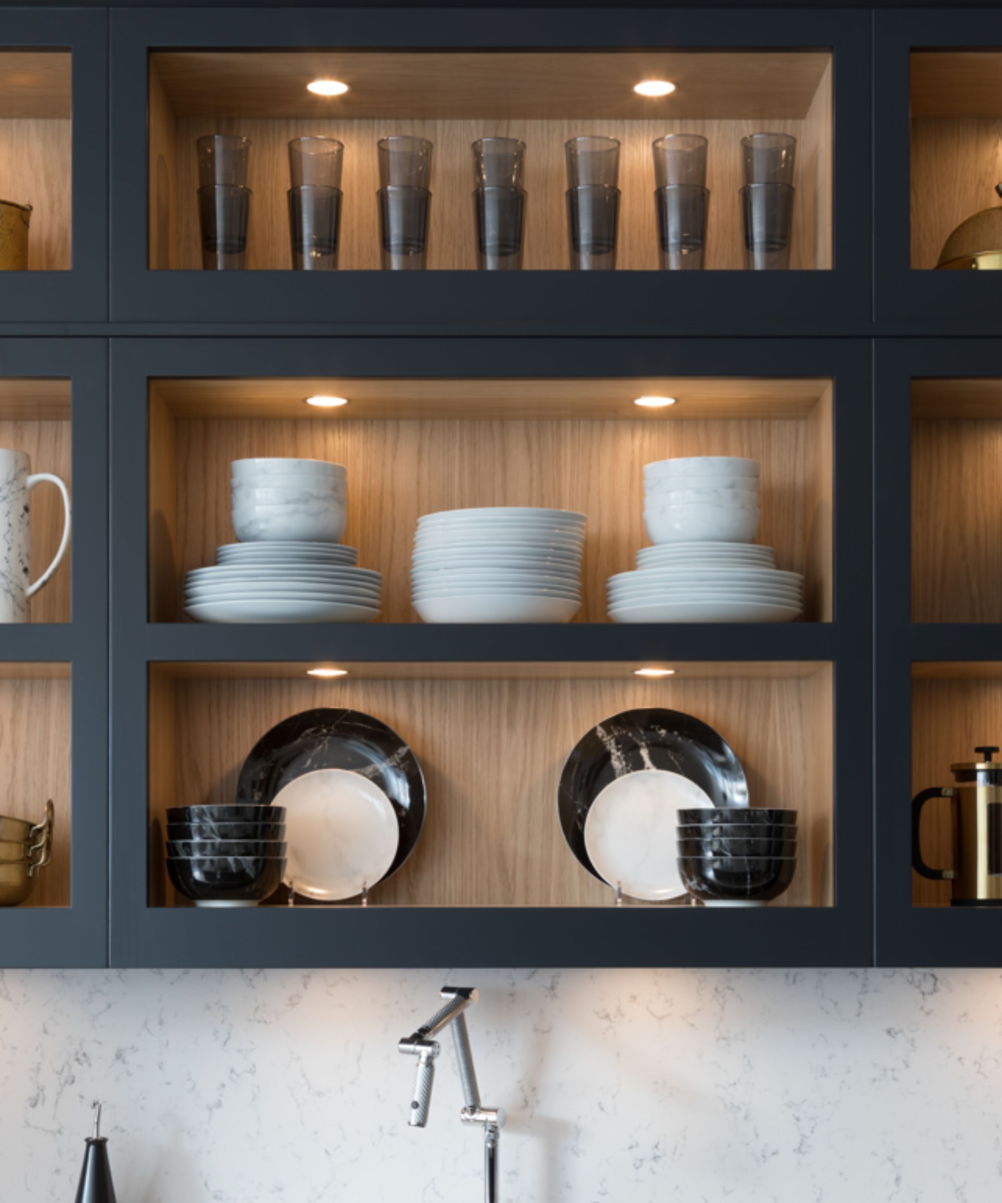 A close-up of open kitchen shelving with integrated warm LED lighting, showing neatly stacked white plates, bowls and clear drinking glasses arranged inside dark blue cabinets with light wood interiors.