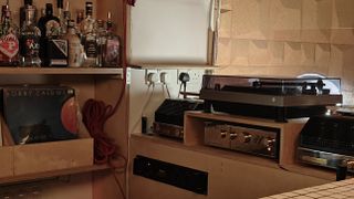 Turntable at the bar top in Behind This Wall. Bottles of alcoholic drinks are lined up to the left, and boxes of vinyl are under the shelf.