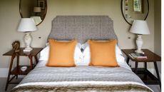 Close up of a double bed with neutral colors of bedding and two propped up plump orange cushions. There are matching wall-mounted brown oval wooden mirrors each side, with classic white lamps and mid-century wooden nightstands
