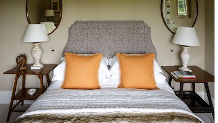 Close up of a double bed with neutral colors of bedding and two propped up plump orange cushions. There are matching wall-mounted brown oval wooden mirrors each side, with classic white lamps and mid-century wooden nightstands