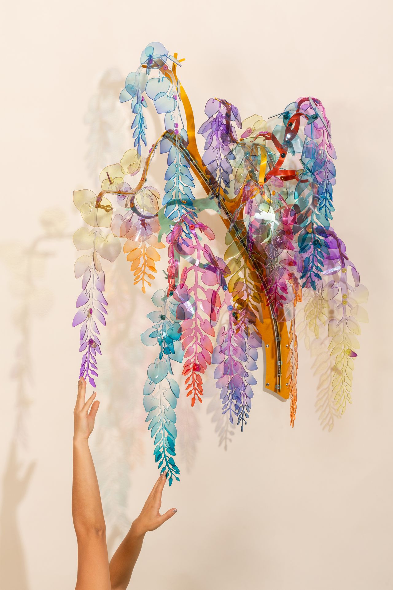 A wisteria-inspired sculptural installation in purple, blue, orange, green, and red sits against a neutral beige background as two human hands reach out to its 'branches'.