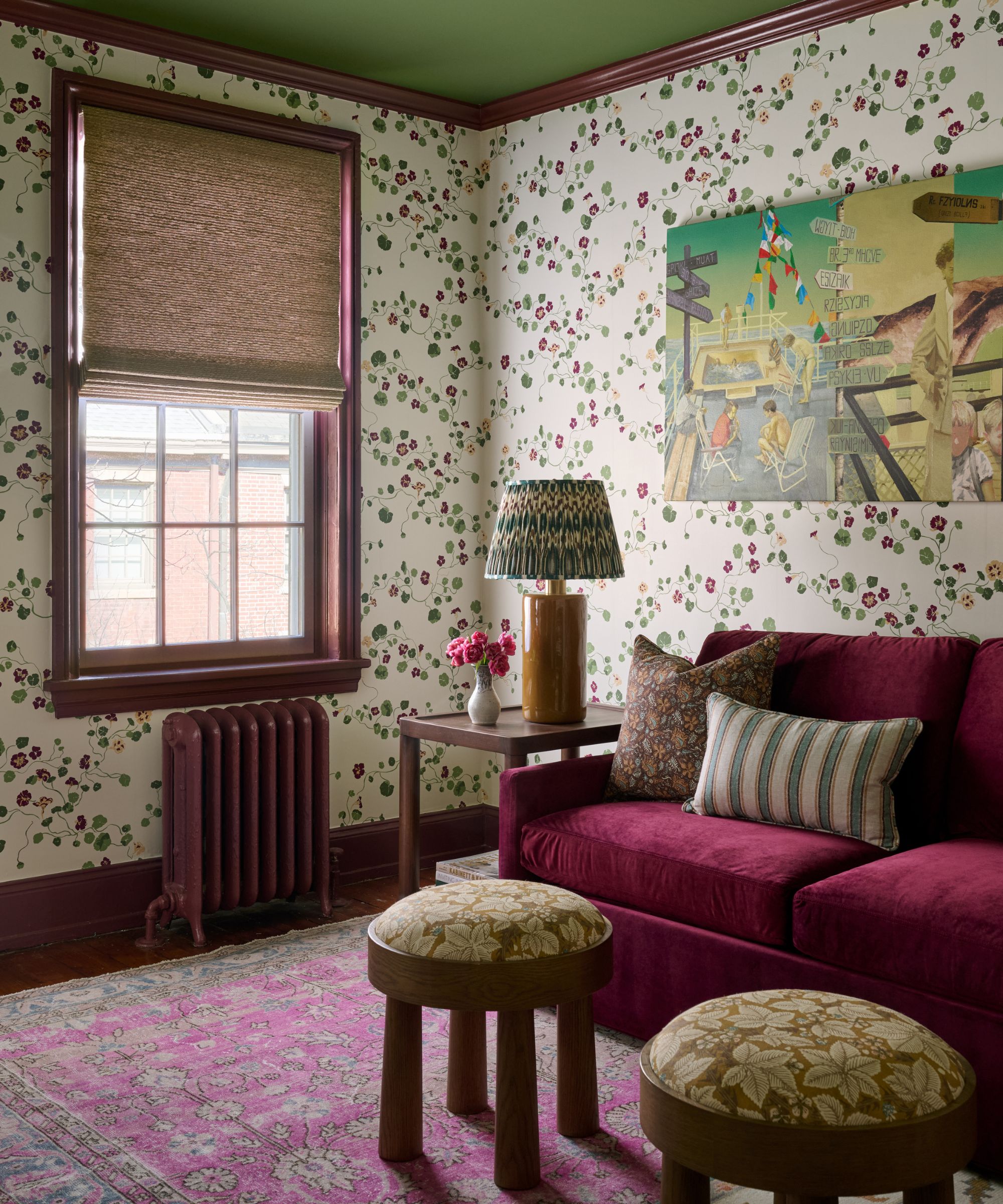 Sitting room corner with floral wallpaper, a burgundy velvet sofa, a pink patterned rug, and two small round wooden stools.