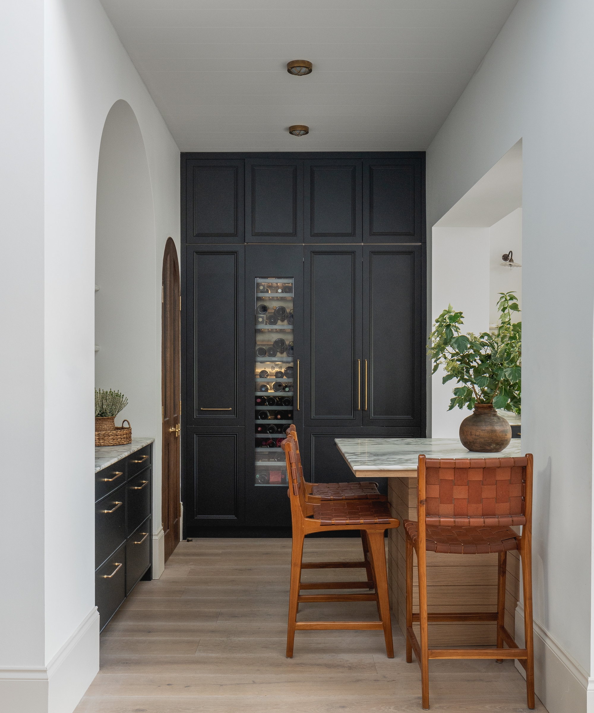 The corner of a kitchen with full height navy cabinetry and an arched wooden door into the pantry