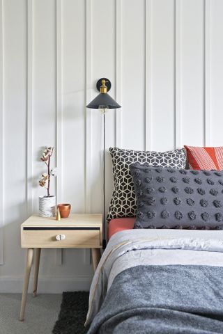 Bed against white vertical panelled wall with light oak bedside table and black wall light