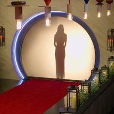 A woman stands behind a circular backlit screen, which opens into a chamber decorated with flowers, Edison bulb lights, candles, and a red carpet, in 'Love is Blind'