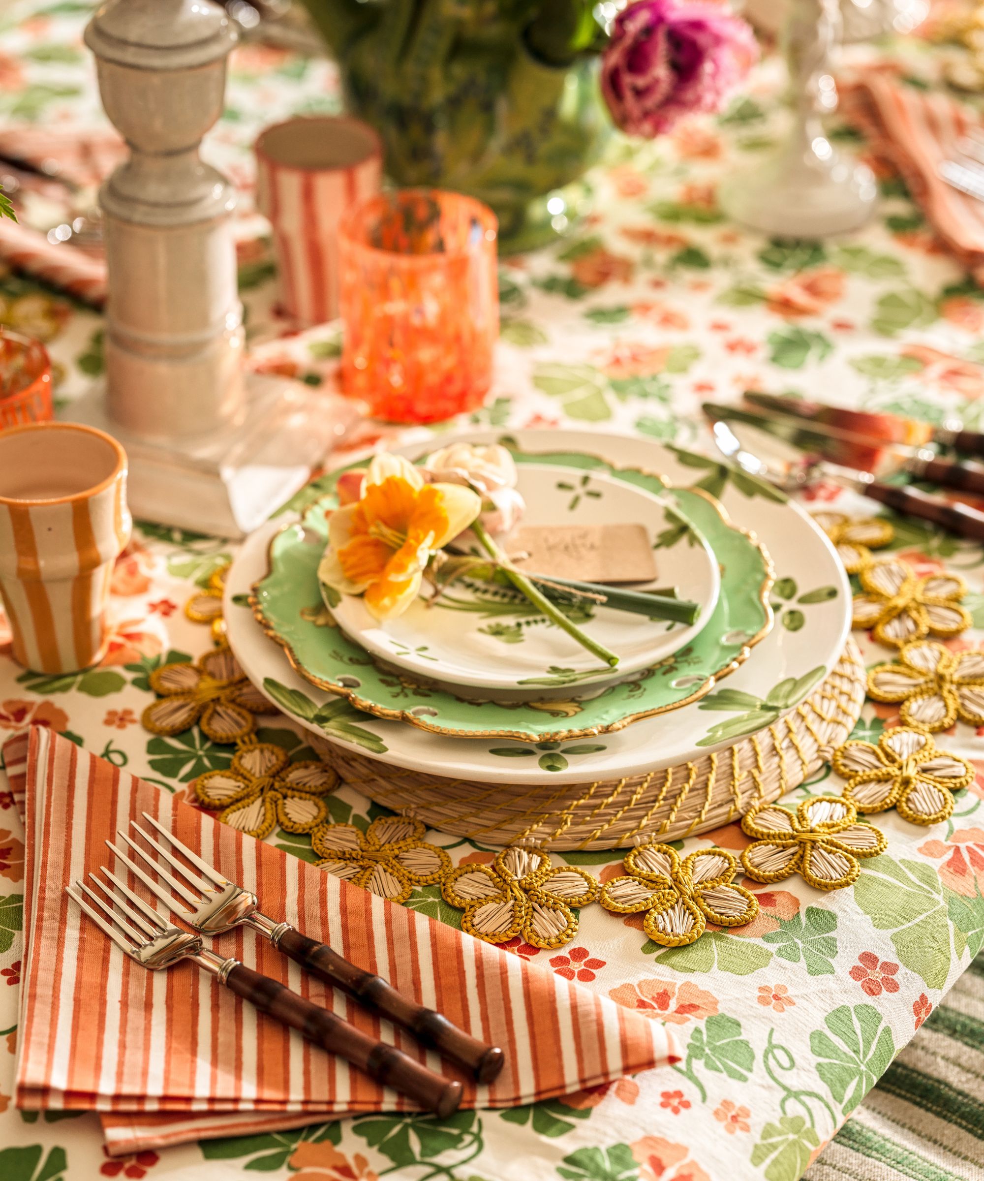 A detailed shot of a place setting featuring bamboo-handled forks on a striped napkin, a green scalloped plate, and a woven floral-shaped placemat on a botanical print tablecloth