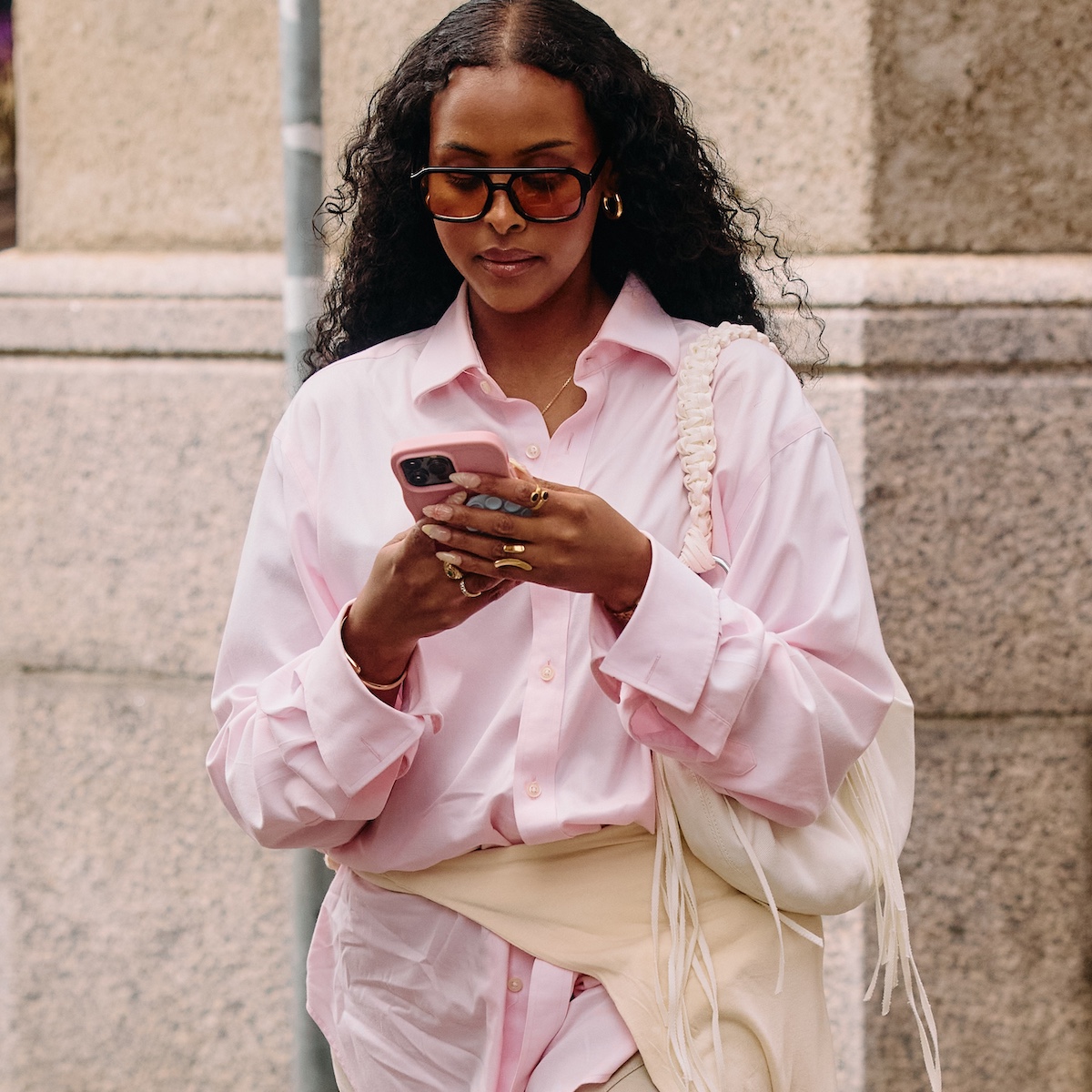 street style shot of a woman looking at her phone wearing aviator sunglasses, a pink button-up shirt, cream shoulder bag, and a sweater tied around her waist