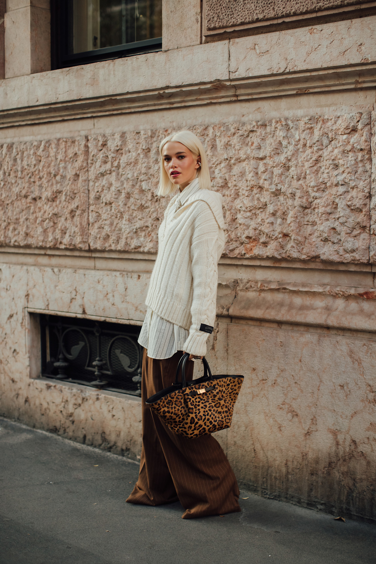 a woman wearing a white cable-knit sweater, striped button-down shirt, brown pants, and a leopard tote bag.