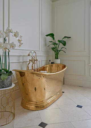 a gold freestanding bath tub in a white bathroom with fresh plants