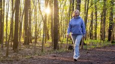 woman in a blue hoodie and jeans walking through a forest with the sun shining through the trees behind her.
