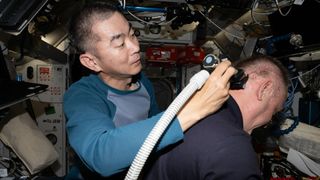 A man holds a razor attached to a hose and slowly shaves another man's head while aboard the ISS
