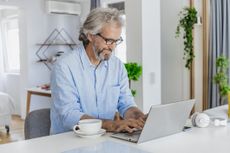 A mature man is using a laptop at home, he is drinking morning coffee and surfing the Net