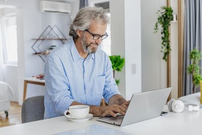 A mature man is using a laptop at home, he is drinking morning coffee and surfing the Net