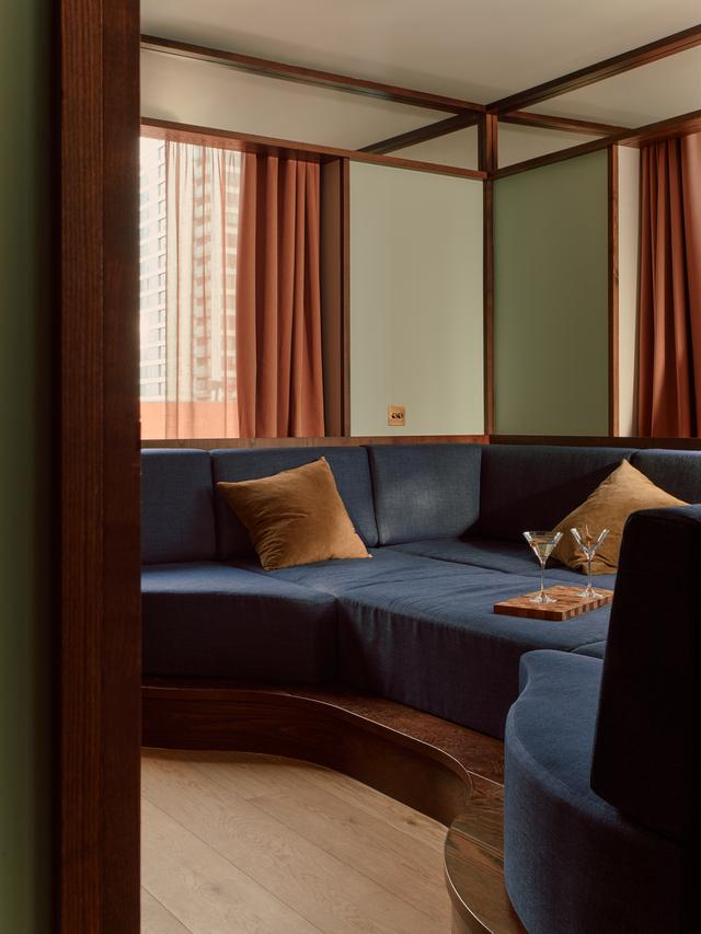 Close up of a muted navy blue cushioned banquette with wooden base and detailing, inserted into a hotel room with wooden floors, terracotta curtains, and green-painted walls.