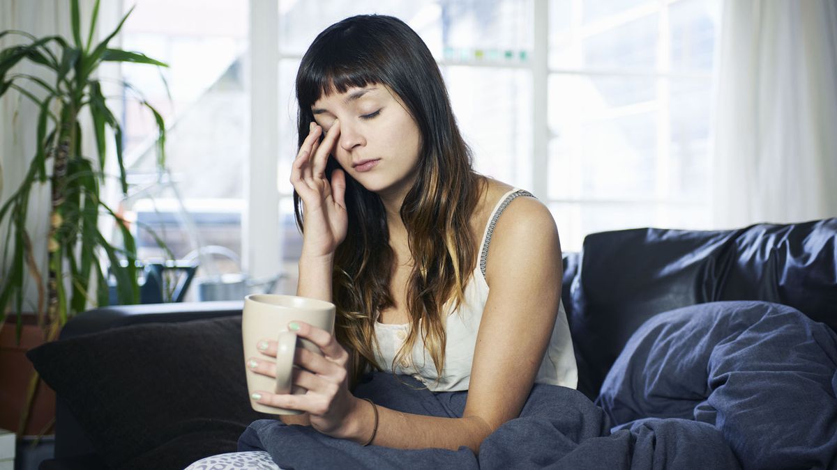 Woman sat up with a duvet wrapped around her, holding a mug and looking tired