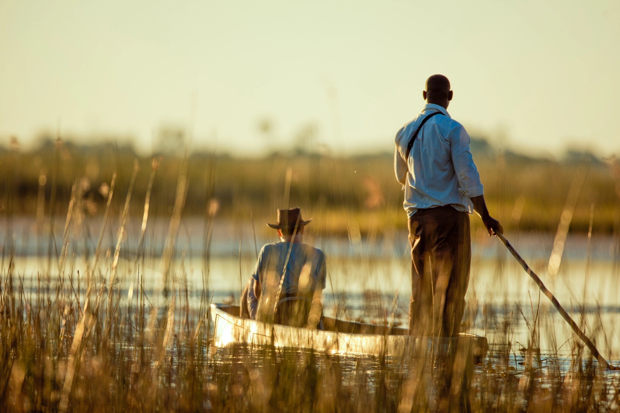 Two men in a canoe at sunset are photographed in a picture, one sitting at its front with a hat, the other, portrayed paddling.
