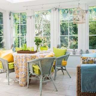 Brightly coloured conservatory with green wicker chairs and table decked in floral tablecloth