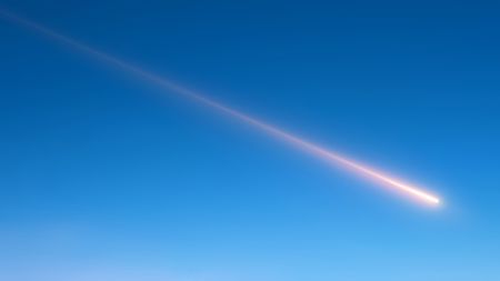 A streak of white moves across the blue sky as a meteor shoots past.