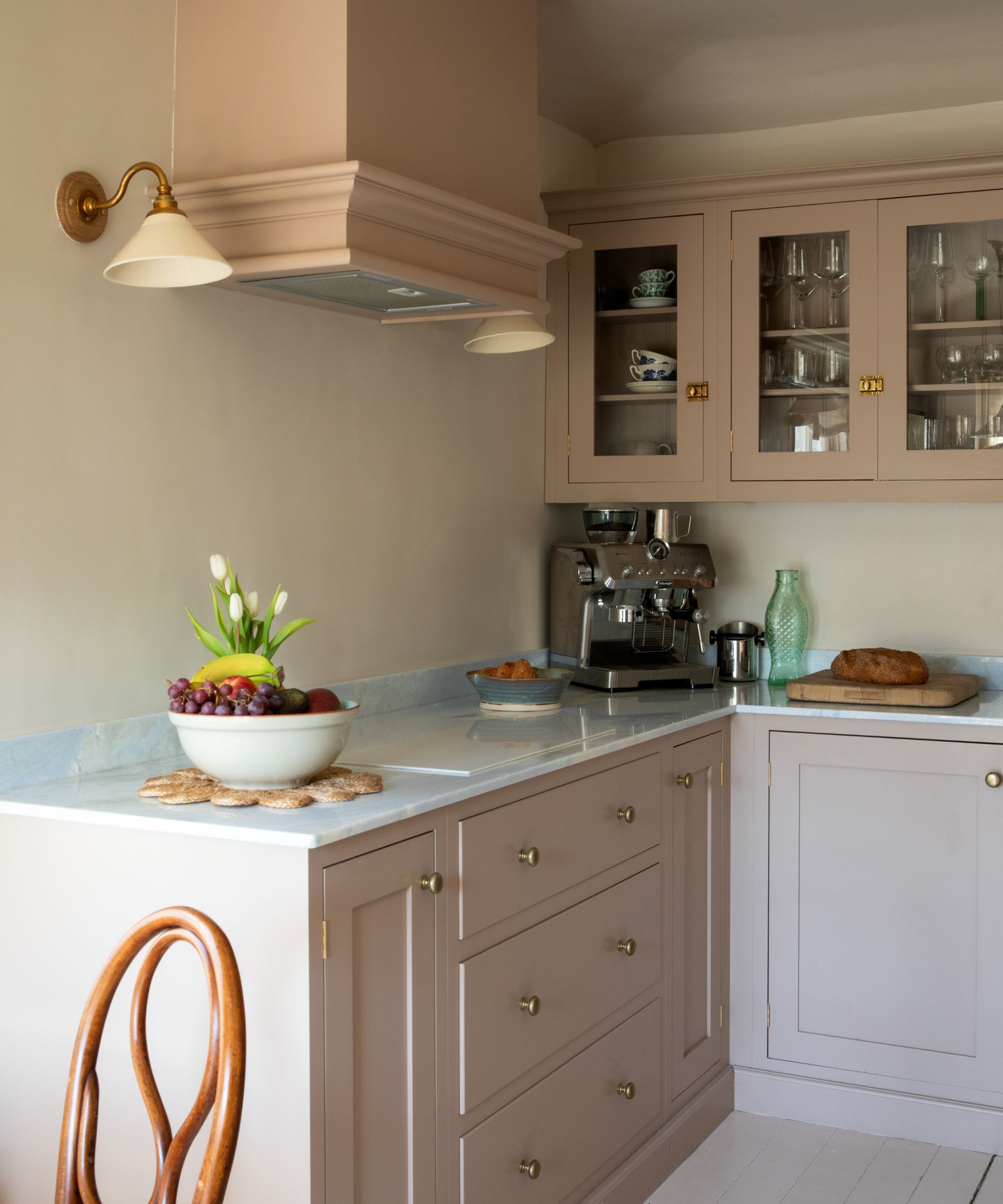 Kitchen with pale pink painted cabinetry, marble countertops, glass-fronted cabinets with glasses and table ware inside, a coffee machine in the corner and gold sconces on the wall