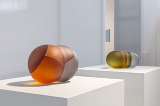 green and orange glass circular objects on a white plinth at the ’Translucency’ exhibition, 8th Tallinn Applied Art Triennial at Kai Art Center
