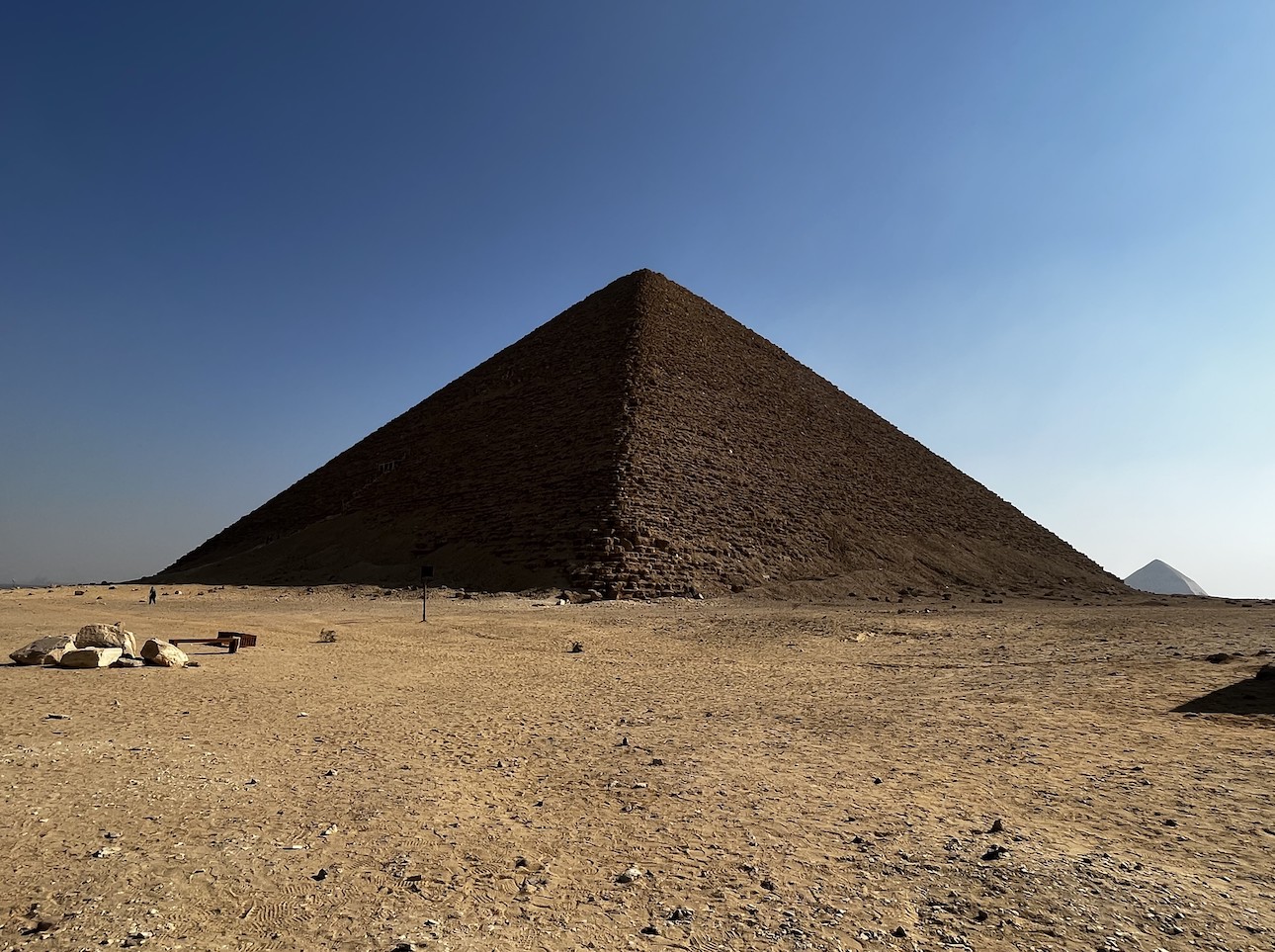 A large pyramid made of stone in the foreground with a smaller one in the background to the right. A few stones are on top of the sand to the left and a small person is in front of the pyramid.