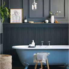 dark panelled wall behind blue roll top bath, wooden flooring and metallic lamp and ornaments.