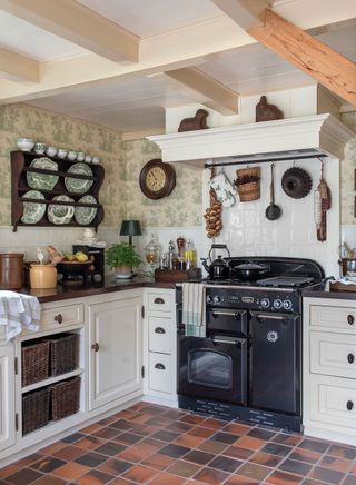 kitchen with black range cooker red tiled floor green toile de Jouy wallpaper and white cabinets vintage plates in rack above