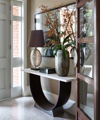 A modern hallway idea with large modern console table with curved underside, topped with table lamp and vase of flowers