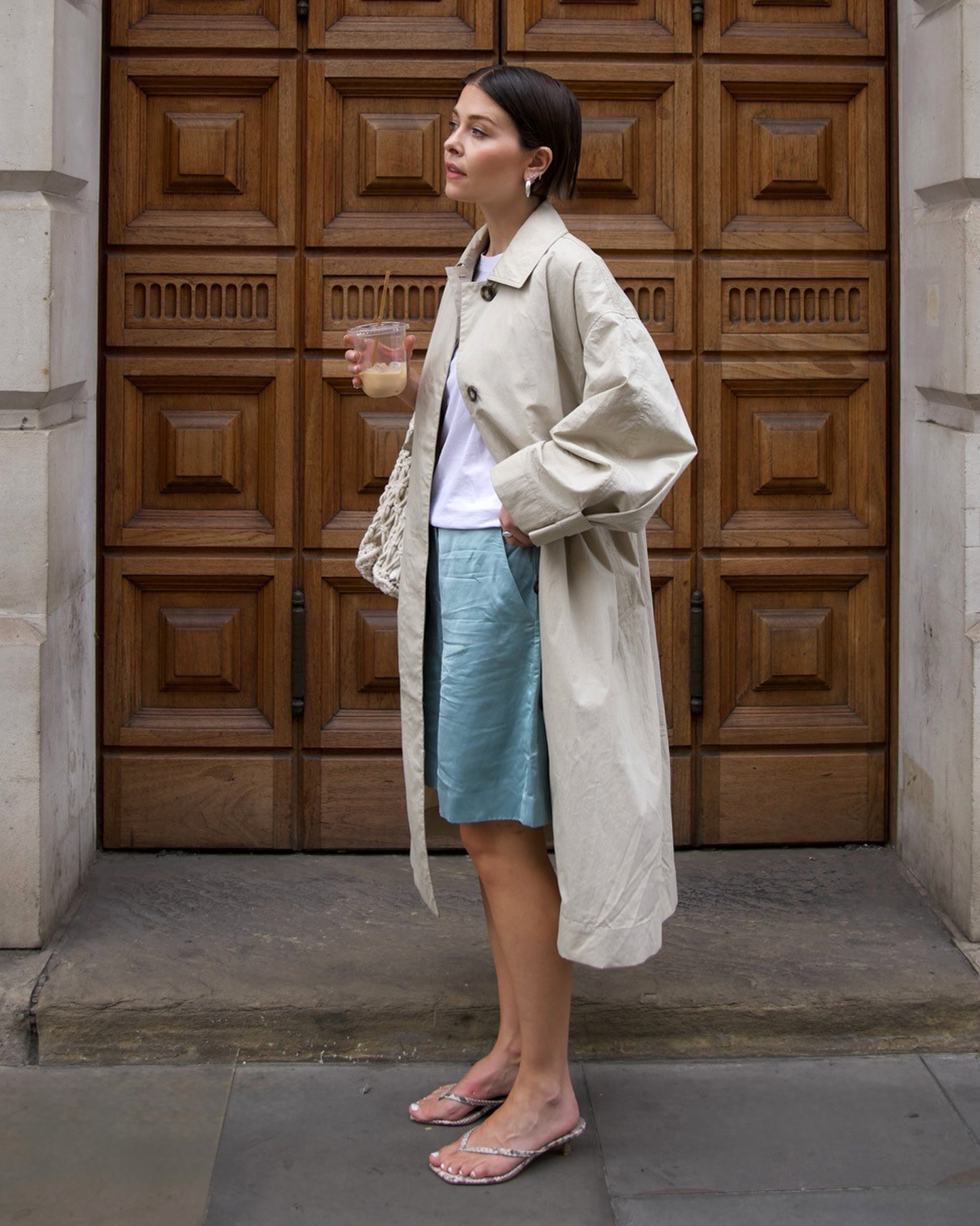 British style influencer Alice Satterthwaite poses in London wearing a mac trench coat, white T- shirt, blue satin Bermuda shorts, snakeskin flip-flop kitten heels