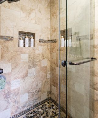 A corner of a terracotta tiled shower with an in-built shelf with three toiletry bottles, black and white stone tiled flooring, and a glass door with a metal handle