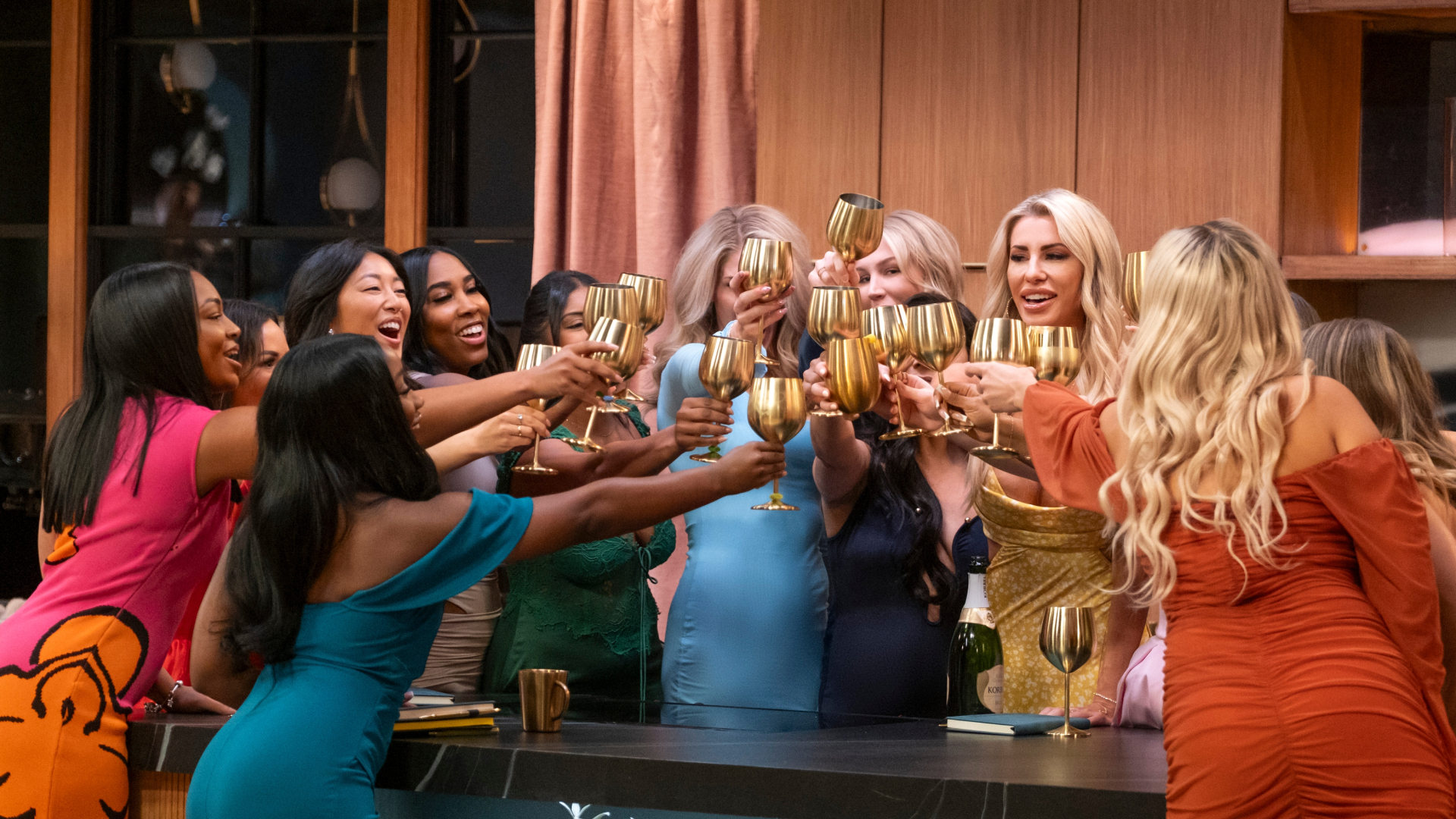 A group of women (l-r: Tyler Lanier, Dynasty Ballard, Emma Betsinger, Bry Thomas, Priyanka Grandhi, Amber Morrison, Christine Hamilton, Elissa Finley, and Rosalyn Ransaw) toast with gold wine glasses as they stand over a kitchen island, in the 'Love is Blind' season 10 premiere.