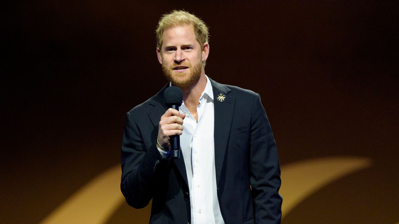 Prince Harry wearing a blue suit speaking into a mic in front of a dark background