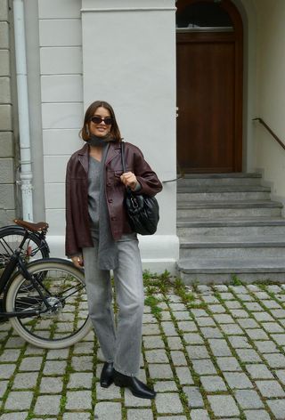 A woman wearing a red leather jacket layered over a gray sweater styled with a gray scarf, gray jeans, black ankle boots, and a black bag.