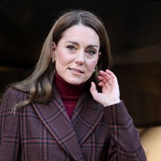 Kate Middleton has long brown wavy hair and wears a brown blazer with jeans, while wearing a tartan overcoat and heels