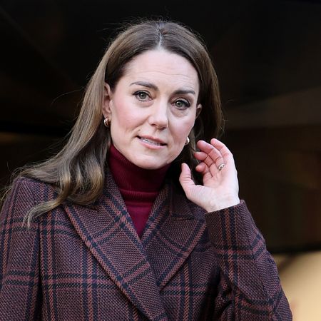 Kate Middleton has long brown wavy hair and wears a brown blazer with jeans, while wearing a tartan overcoat and heels