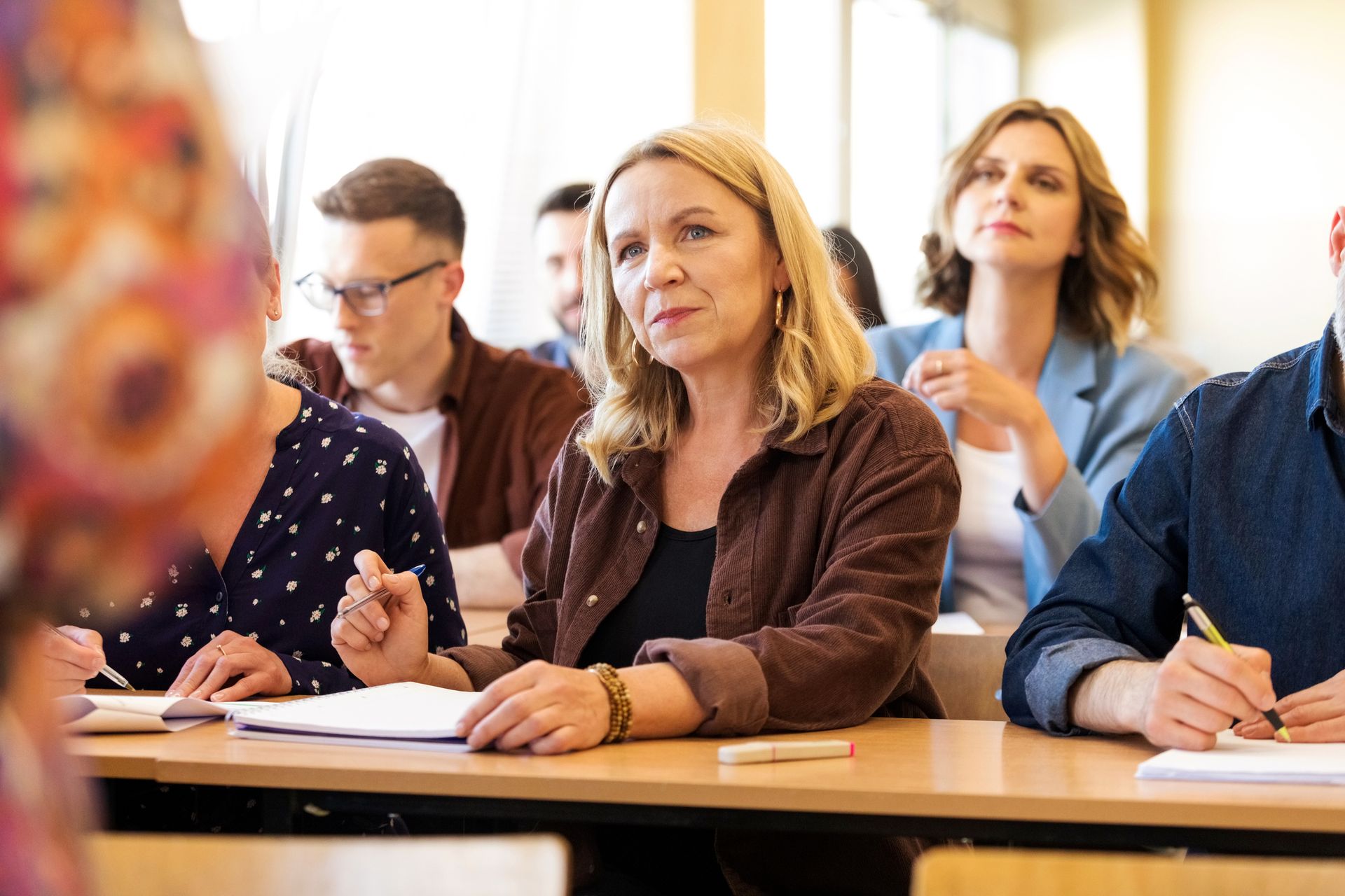 Group of adult female and male students sitting at the desks in the classroom, looking away and listening.