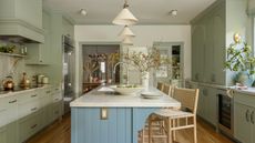 A kitchen with sage green cabinets, a large island with a blue paneled base and a light countertop, featuring woven bar stools and an open view to a dining room
