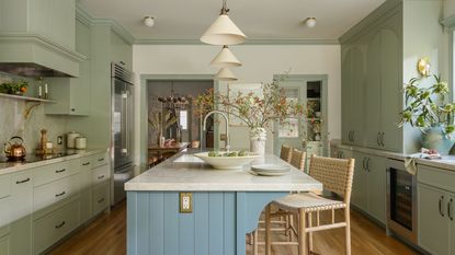 A kitchen with sage green cabinets, a large island with a blue paneled base and a light countertop, featuring woven bar stools and an open view to a dining room