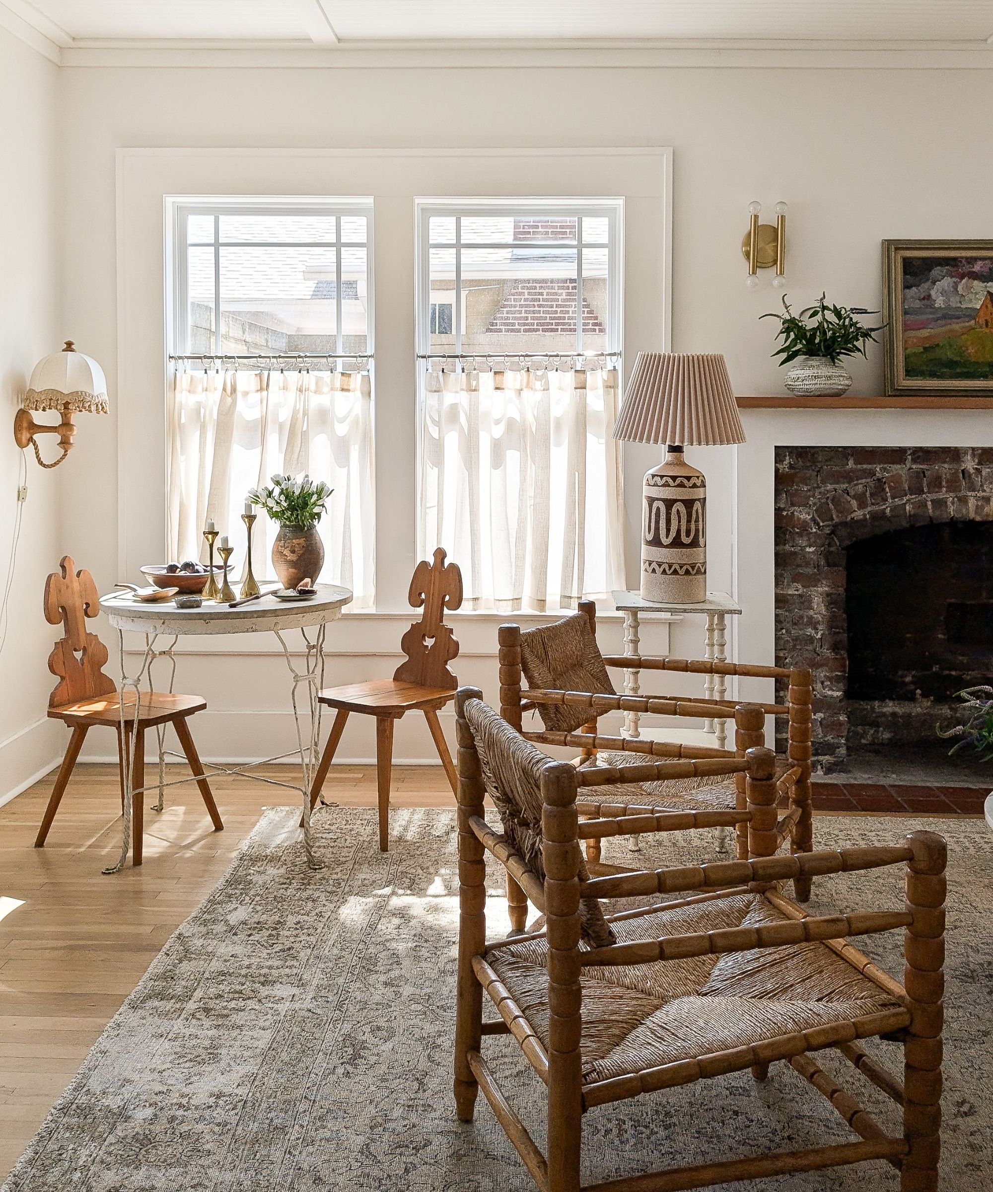 A sun-drenched living area with white walls, cafe curtains, and a brick fireplace. The room is furnished with a wire-base bistro table, decorative carved wood chairs, and woven spindle armchairs on a faded vintage rug