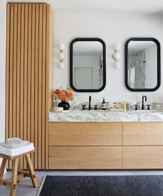 Contemporary bathroom with a wooden vanity topped with a marble countertop. White walls are decorated with black framed mirrors and round wall lights